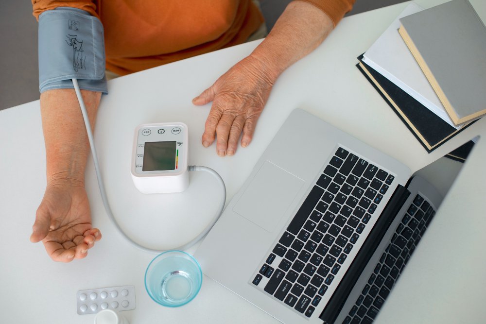 A woman measuring their blood pressure with an automatic bp appratus. She is sitting on a chair and the right hand is resting on the table.