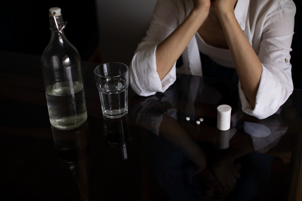 A girl with her hands covering her mouth. An alcohol bottle, glass, and pills are seen on the table