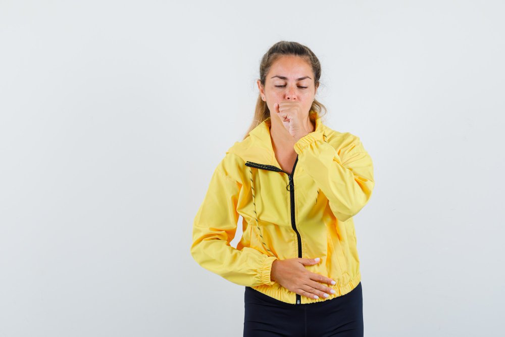 Blonde woman coughing in yellow bomber jacket and black pants and looking hurt